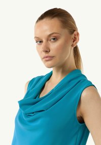 Turkusowy top z dekoltem w formie cowl, wykonany z miękkiej, gładkiej tkaniny i bez rękawów. Drapowanie tworzy eleganckie fałdy w okolicy dekoltu.