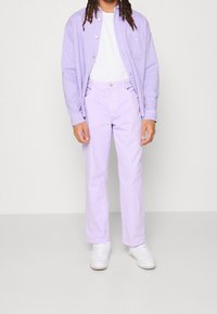 Světle fialová košile na zapínání přes bílým tričkem, doplněná světle fialovými kalhotami a bílými teniskami. Košile má náprsní kapsu.