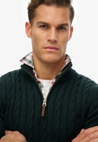 Pull vert à maille torsadée avec col zippé, porté sur une chemise à carreaux. Les détails remarquables incluent un tire-zip en cuir et un tissu texturé.