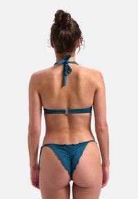 Tyrkysová plavková sada s halter krkem a zavazováním, nařasenými detaily a minimálním pokrytím na spodní části s volánkovými okraji.