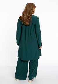Langes, fließendes teal-farbenes Outfit mit einem plissierten Rücken, langen Ärmeln und lockeren Hosen. Glatte Stofftextur mit einer entspannten Silhouette.