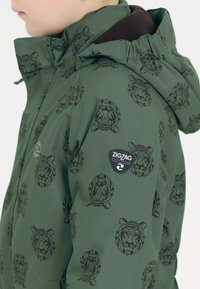 Groene jacket met een zwart geometrisch leeuwpatroon. Het heeft een capuchon, verstelbare manchetten en een prominent zwart 'ZIGZAG'-logo patch.