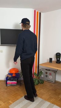 Black sweater and pants; orange mushroom-shaped lamp on a blue table; gray speaker; potted plant; striped wall; wooden floor.