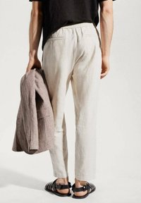 Pantaloni in lino beige chiaro con vita elastica, abbinati a un blazer in lino grigio drappeggiato su un braccio e sandali neri.
