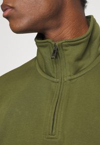 Gros plan sur la partie inférieure du visage et le cou d'une personne portant un sweat-shirt vert olive à demi-fermeture éclair avec le col partiellement ouvert.