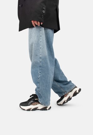 Person iført løstsiddende lyseblå jeans, sort blazer og sorte og hvide sneakers med leopardprintdetaljer, stående på en hvid baggrund.