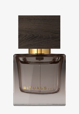 Vierkante glazen parfumfles met rokerig verloop en gouden "RITUALS" tekst, bovenop een grote donkere houten rechthoekige dop en gouden kraag.