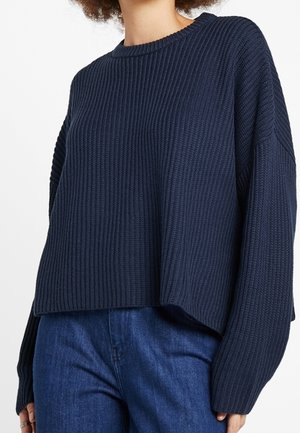Kobieta w luźnym, ciemnoniebieskim swetrze z prążkowanego dzianiny, zestawionym ze średnio niebieskimi dżinsami, widziana od szyi do bioder.