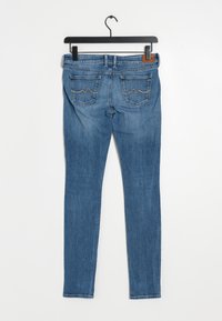 Blå denim skinny jeans hängande på en svart galge, med två bakfickor med brunt kantband och subtila sömnadsdetaljer.