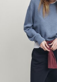 Blue crewneck sweater with long sleeves, textured fabric, worn over a white top, and a maroon garment tied around the waist. Black pants.
