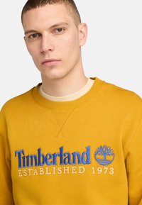 Gul sweatshirt i bomull med blå broderad logotyp och texten "Timberland ETABLERAD 1973" på framsidan. Rund halsringning.