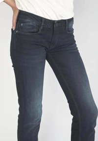 Jean en denim bleu foncé avec une coupe slim, présentant des poches avant, une fermeture à bouton sur le devant, et un léger décoloration sur les jambes.