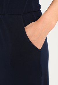 Marinblå hoppdräkt med en slät yta, som har en sidficka där en hand är placerad, vilket framhäver dess praktiska design.