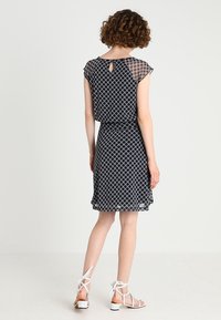 Robe noire avec motif géométrique blanc, manches courtes à capuchon, taille ajustée et détail du dos en forme de clé. Portez-la avec des sandales blanches à lanières.