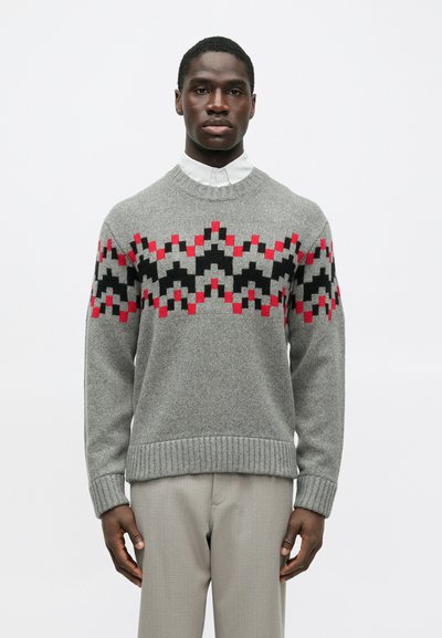 Pull gris avec un motif géométrique en noir, blanc et rouge ; ourlet et poignets côtelés ; col rond ; associé à un pantalon de couleur claire.