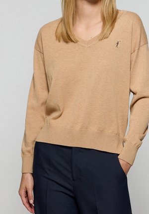 Beżowy sweter z dekoltem w serek z małym haftowanym logo na piersi, noszony z ciemnogranatowymi spodniami; lewa ręka osoby w kieszeni, prawa ręka swobodnie opada.