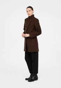 Cappotto in misto lana marrone con colletto alto, taglio dritto, tasche e finiture in oro. Abbinato a pantaloni neri a gamba larga e stivaletti neri.