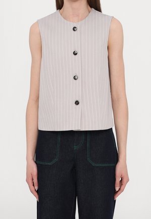 Bezrękawnik w jasnoszarym kolorze w prążki, z czterema czarnymi guzikami z przodu, noszony z ciemnymi spodniami z wysokim stanem z zielonym przeszyciem.