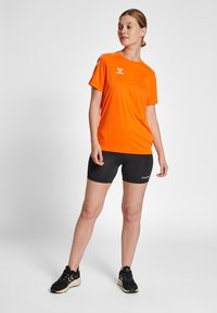 Ljusorange kortärmad träningsskjorta med logotyp, tillsammans med svarta kompressionsshorts. Modellen bär svarta sneakers och står mot en neutral bakgrund.