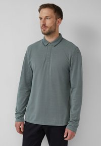 Homme avec une barbe légère portant un polo à manches longues vert discret et un pantalon noir, se tenant devant un fond gris clair uniforme.