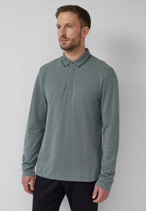 Man met een lichte baard, gekleed in een muted groene polo met lange mouwen en zwarte broek, staande tegen een effen lichtgrijze achtergrond.