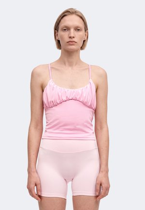 Femme aux cheveux blonds courts portant un haut caraco froncé rose et un short de vélo taille haute assorti, debout devant un fond gris uni.