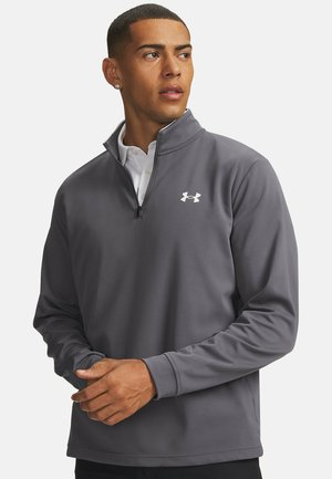 Junger Mann trägt einen grauen Viertel-Reißverschluss-Pullover mit weißem Logo, schaut zur Seite und hat die Hände vor einem schlichten Hintergrund verschränkt.