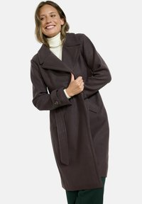 Cappotto di lana marrone con colletto, tasche laterali e cintura. Il cappotto ha una vestibilità rilassata e una superficie liscia, indossato sopra un dolcevita crema.