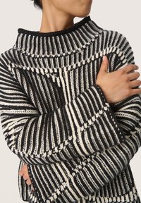 Gestreifter Pullover in Schwarz und Weiß mit hohem Kragen, der ein strukturiertes Strickmuster und weite Ärmel aufweist. Kontrastierendes Garn schafft Tiefe.