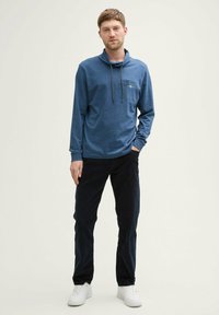 Niebieski długi rękaw sweter z wysokim kołnierzem, sznurkiem i przednią kieszenią. Zestawiony z ciemnymi spodniami i białymi trampkami, w pomieszczeniu.