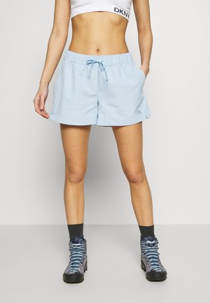 Femme portant un short bleu clair avec cordon, un soutien-gorge de sport blanc, des chaussettes foncées et des chaussures de randonnée grises, debout devant un fond uni.