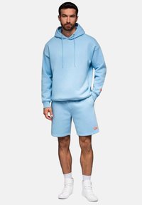 Lichtblauwe hoodie en bijpassende shorts van zachte stof, met een capuchon, kangoeroezak en logoaccent op de shorts. Witte sneakers maken de look compleet.