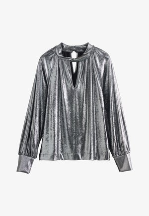 Femme portant un chemisier argenté brillant à manches longues avec un col en forme de goutte, un pantalon noir à jambes larges et des sandales noires à talons, debout avec une main dans la poche.