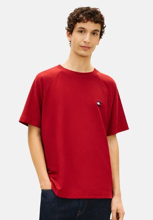 Joven con cabello rizado, vestido con una camisa roja de manga corta y jeans oscuros, de pie con una mano en el bolsillo contra un fondo liso.