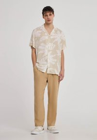 Camicia a maniche corte, di colore chiaro, con motivi a foglia bianchi, abbinata a pantaloni beige a vestibilità comoda e sneakers bianche.