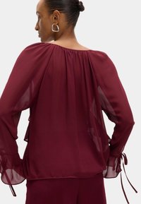 Bordeaux sheer blouse met een geplooide halslijn, vloeiende mouwen en zijstrikjes. Lichtgewicht stof met een zachte textuur en elegante val.