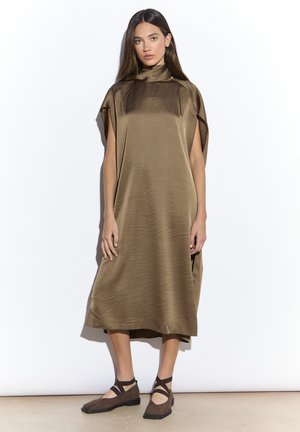 Mujer de pie contra un fondo blanco, vestida con un vestido suelto de raso verde oliva de longitud media, de cuello alto, y zapatos de ballet marrones con tiras al tobillo.