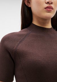 Κοντινό πλάνο ενός ατόμου που φοράει καφέ μπλούζα με στενή εφαρμογή, κοντά μανίκια ριμπ, ψηλό λαιμό και εμφανείς ραφές.