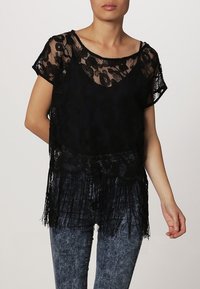 Svart spetsblus med korta ärmar, blommönster och fransdetaljer vid nederkanten, ihop med blå texturerade jeans.
