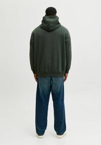 Sudadera verde oscuro con una gran capucha suave, combinada con jeans azules claros y holgados. La tela parece acogedora y sin costuras, con puños de canalé.