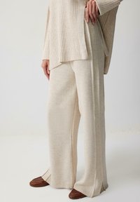 Pantaloni din tricot ribbed bej, cu deschideri pe laterale la tiv, asortați cu un top texturat corespunzător, purtați cu pantofi maro slip-on.