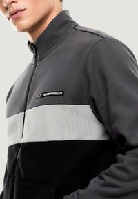 Chaqueta con cremallera en negro, gris y blanco, con un alto cuello. Presenta un diseño bicolor y un logo bordado en el pecho. Material suave.