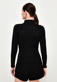 Schwarzes Langarmoberteil mit hohem Hals und gerippter Textur, mit vertikalen Linien und einer figurnahen Silhouette, kombiniert mit schwarzen Shorts.