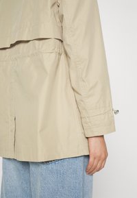 Beige lättviktig jacka med lagerdesign, synlig sömnad och elastisk drapering vid ärmarna. Bärs över ljusblå denimbyxor.