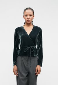 Tummanvihreä samettinen kietoutuva paita, jossa on pitkät hihat ja sidottava vyötärö. Ominaisuutena syvä V-kaula-aukko ja sileä pinta. Yhdistetty korkeavyötäröisten harmaiden housujen kanssa.