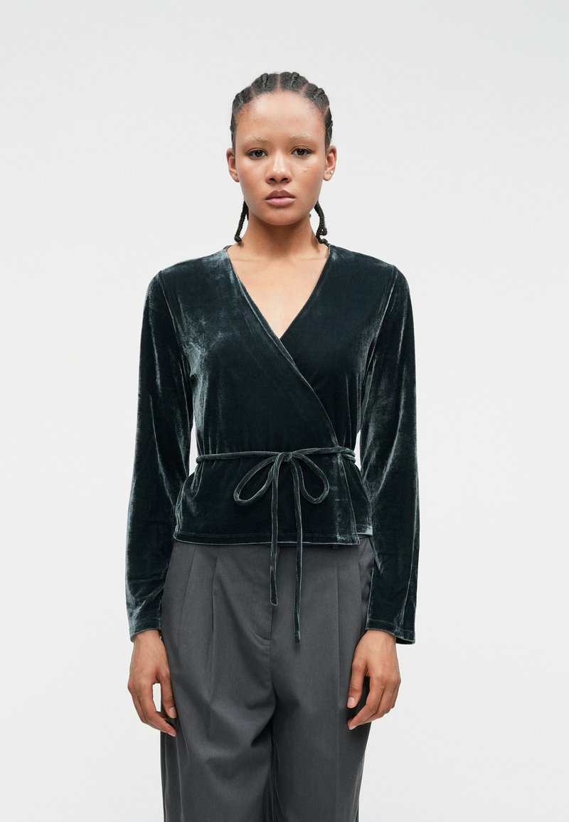 Tummanvihreä samettinen kietoutuva paita, jossa on pitkät hihat ja sidottava vyötärö. Ominaisuutena syvä V-kaula-aukko ja sileä pinta. Yhdistetty korkeavyötäröisten harmaiden housujen kanssa.