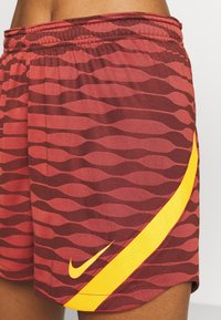 Rote und burgunderfarbene Shorts mit Wellenmuster. Mit einem gelben Nike-Swoosh, elastischem Bund und strukturiertem Stoff.