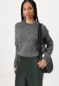 Maglione grigio a maglia con colletto rotondo, abbinato a pantaloni in velluto a coste verdi. Una borsa a tracolla nera presenta un marchio impresso. Dettagli del materiale testurizzato visibili.