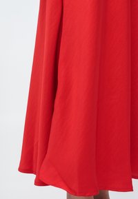 Czerwony, płynący materiał sukienki o gładkiej fakturze. Linia dołu delikatnie się zaokrągla, tworząc lekko rozkloszowaną sylwetkę. Brak widocznych wzorów czy elementów metalowych.