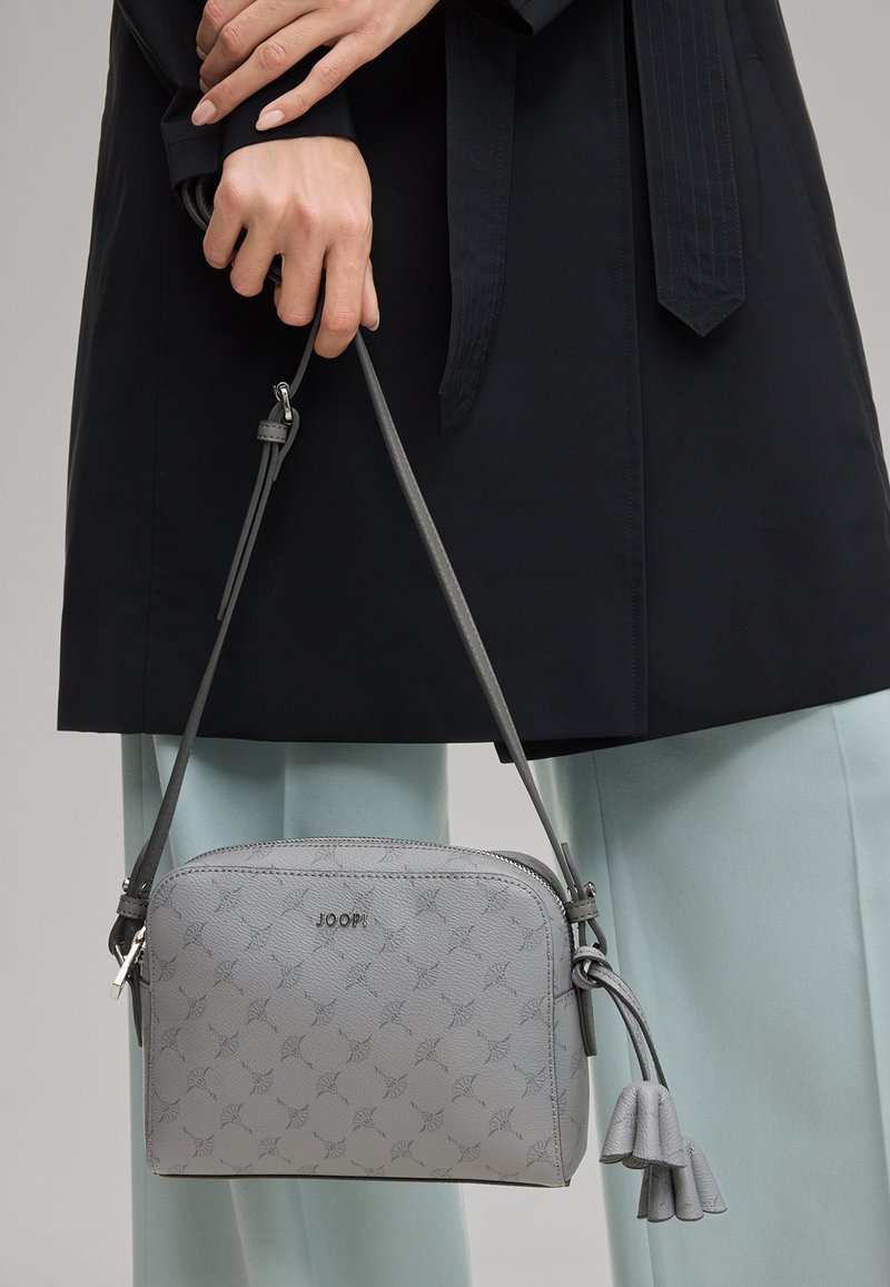 Hænder der holder en grå crossbody-taske med mønster og kvastdetalje, båret med en sort frakke og lyseblå bukser.